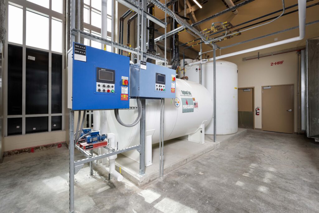 Large white fuel storage tanks in a mechanical room with blue control panels mounted on a metal frame.