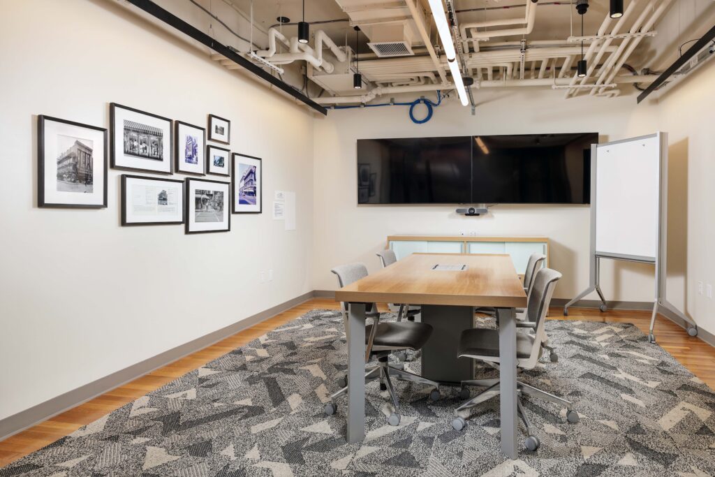 A modern conference room with a wooden table, four chairs, a wall-mounted television, a whiteboard, and a gallery of framed black-and-white photos on the wall.