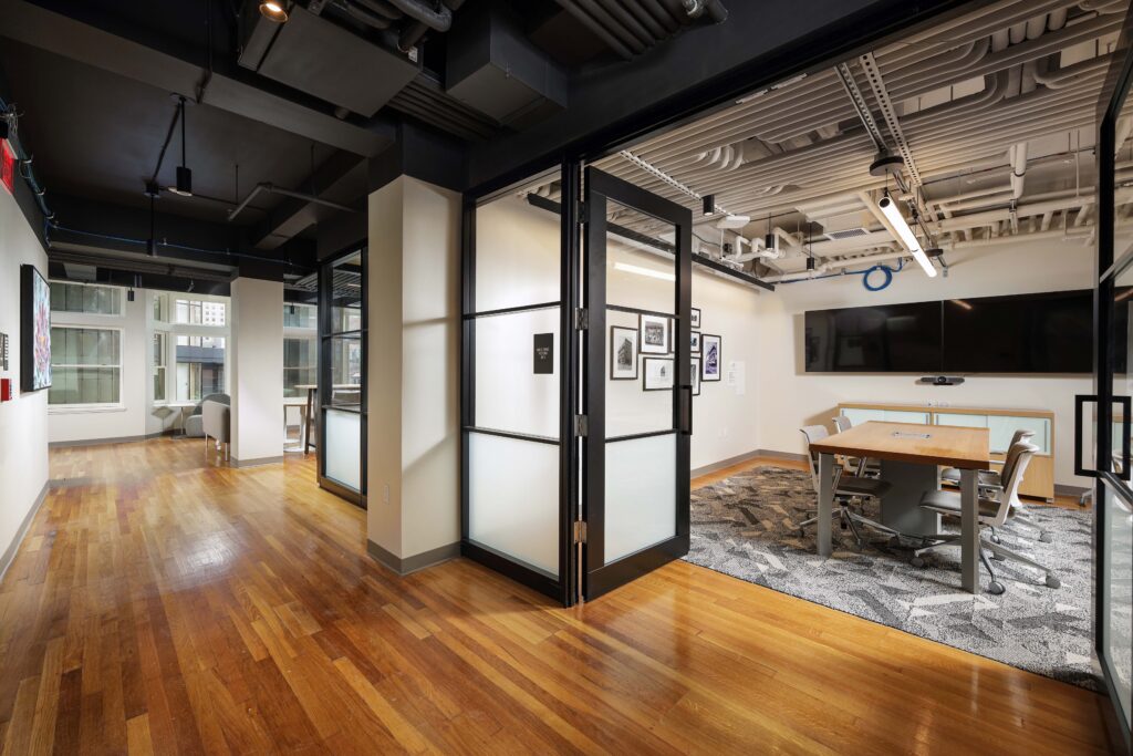 Modern office space featuring a conference room with glass doors, a wooden table, and chairs on a patterned rug, situated next to a hallway with hardwood floors.