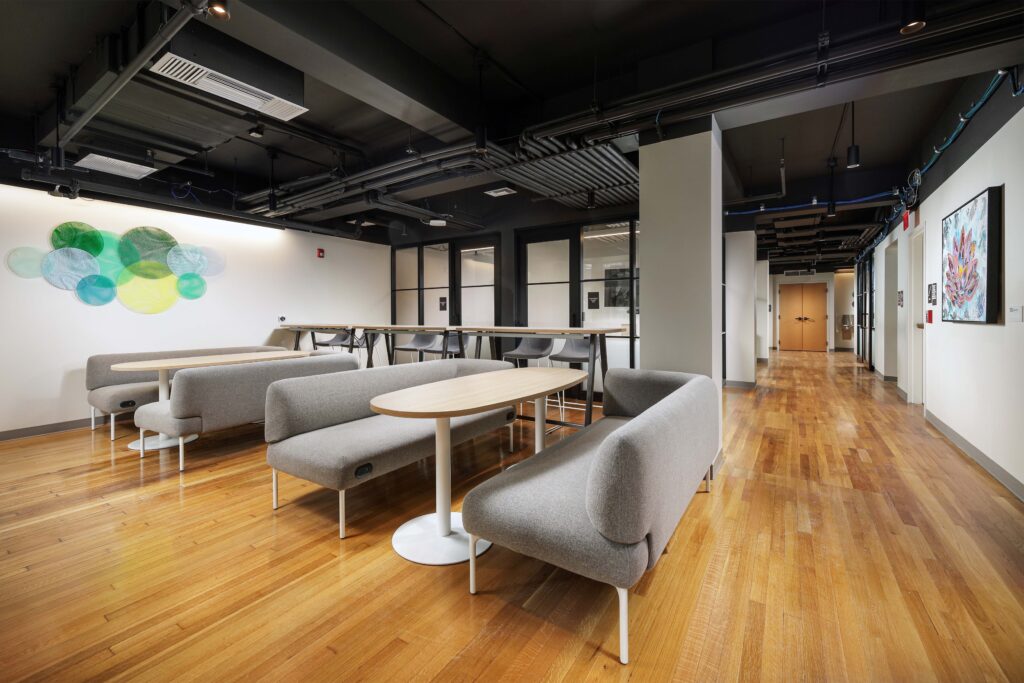 Modern office lounge with gray curved benches, wooden tables, and hardwood floors.