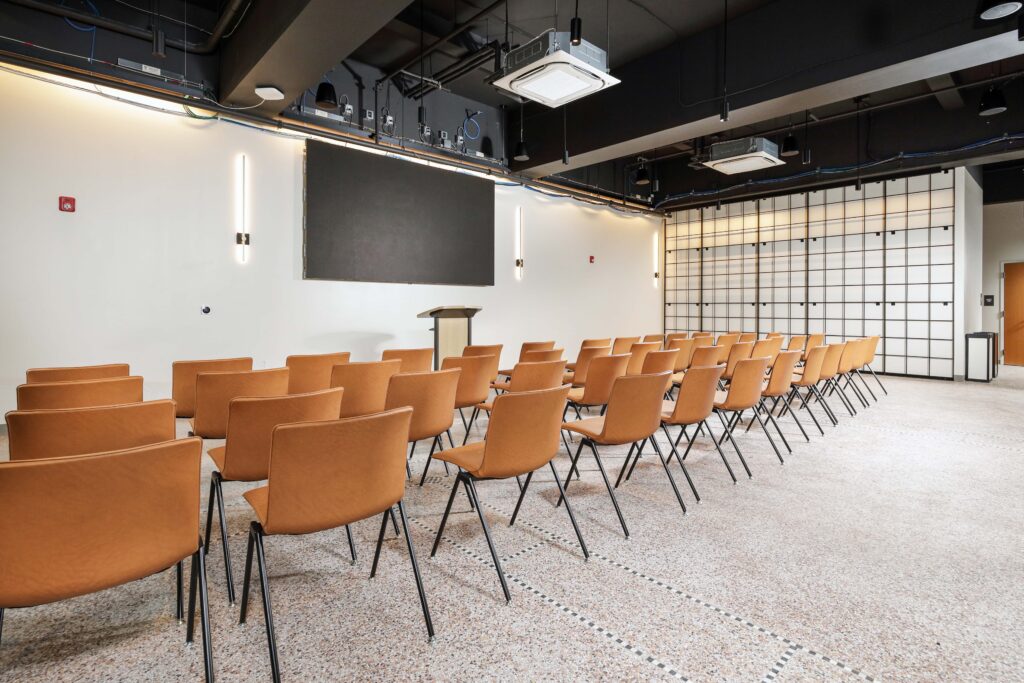 Rows of orange chairs facing a podium and a large screen in a modern conference room.