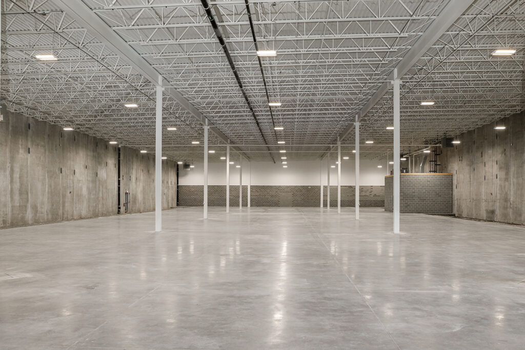 Empty industrial warehouse with a polished concrete floor, concrete walls, and an exposed ceiling structure with overhead lighting.
