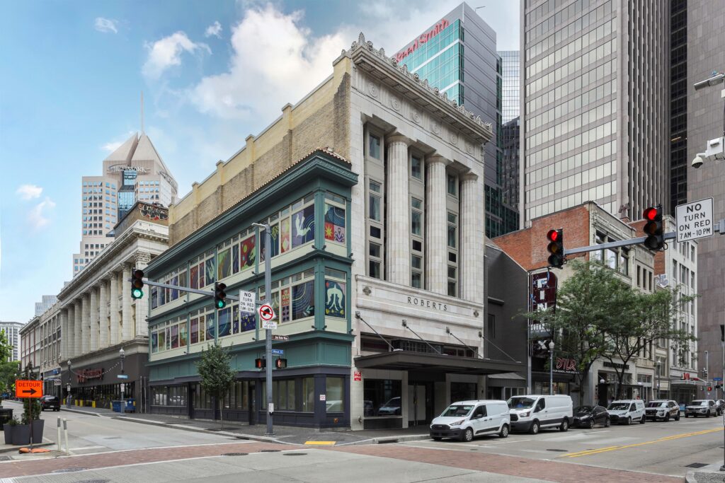 A corner view of the Roberts building in a city, featuring a cream-colored stone facade with columns next to a teal building with decorative stained glass windows.