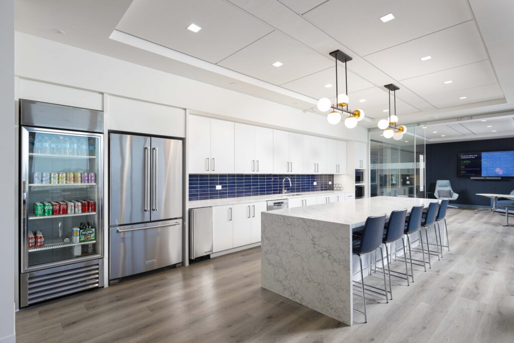 A modern office kitchen with white cabinets, a marble-topped island with five stools, a refrigerator, and a glass-fronted beverage cooler.