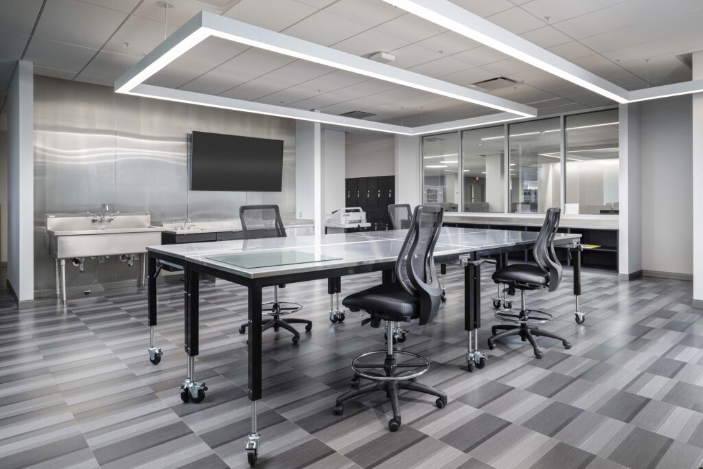 A laboratory-style room with a long metal table on casters, three ergonomic office chairs, a wall-mounted flat-screen monitor, and a stainless steel sink.