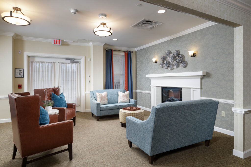 A seating area with two brown leather armchairs, two blue sofas, a round ottoman, and a fireplace with decorative wall art above it.