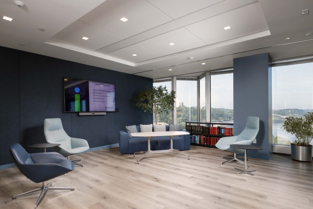 A modern office lounge featuring a blue sofa, light blue armchairs, a coffee table, and a wall-mounted monitor displaying corporate information, with floor-to-ceiling windows overlooking a river view.