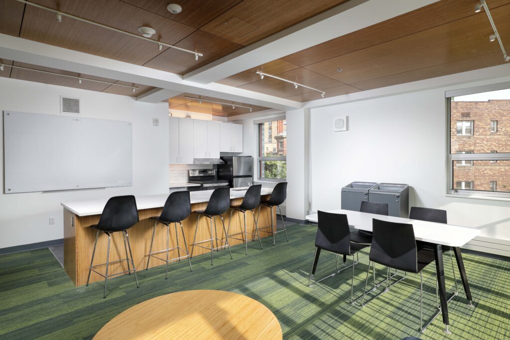 A modern office breakroom with a kitchen counter, bar stools, a dining table with chairs, a whiteboard, and green carpet.