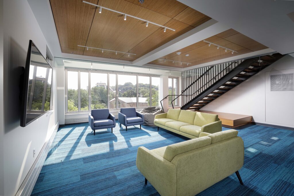 Modern office lounge with two blue armchairs, two green sofas, and a beanbag chair on blue carpet, featuring a wood-paneled ceiling and an open staircase.