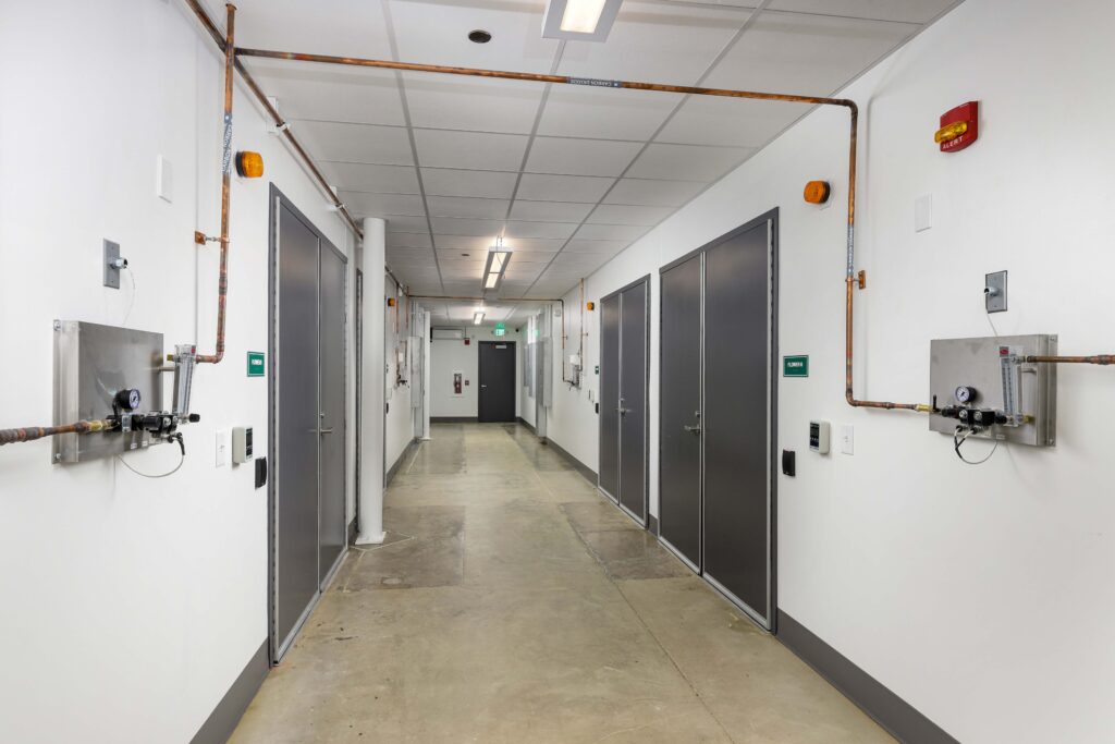 A long hallway with gray doors on both sides, industrial plumbing along the ceiling, and gas monitoring equipment mounted on the walls.