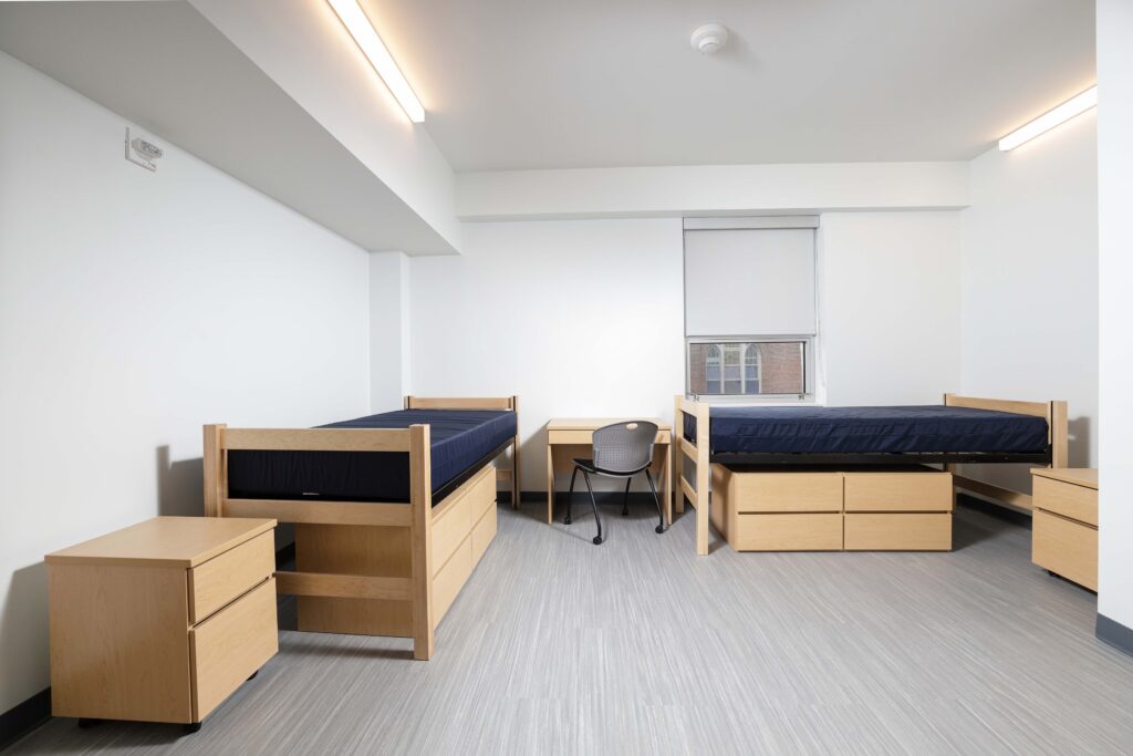 A furnished dorm room with two beds, two dressers, and a desk with a chair.
