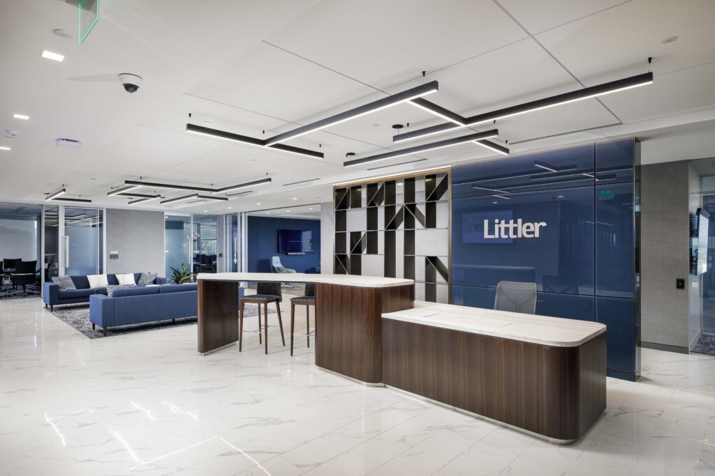 Modern office reception area with a wood and white marble front desk, a blue feature wall with the Littler logo, and an open lounge area in the background.