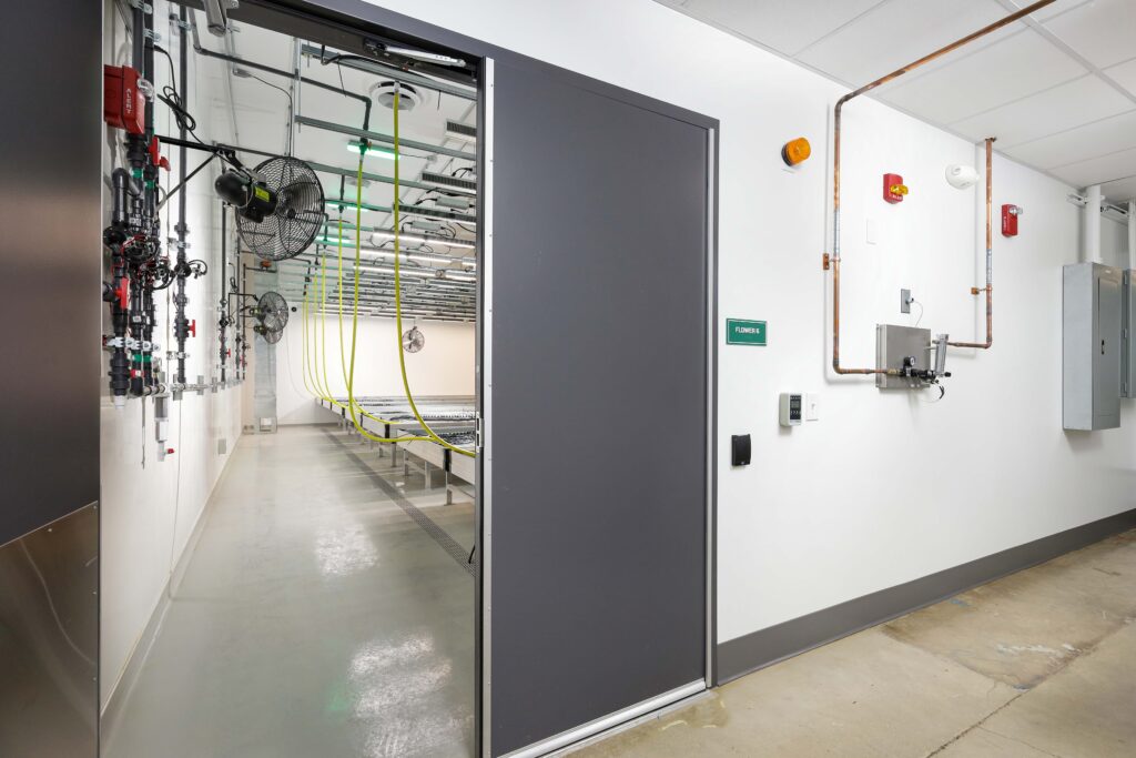 View through an open doorway into an indoor agricultural facility with metal tables, hanging equipment, and yellow hoses.