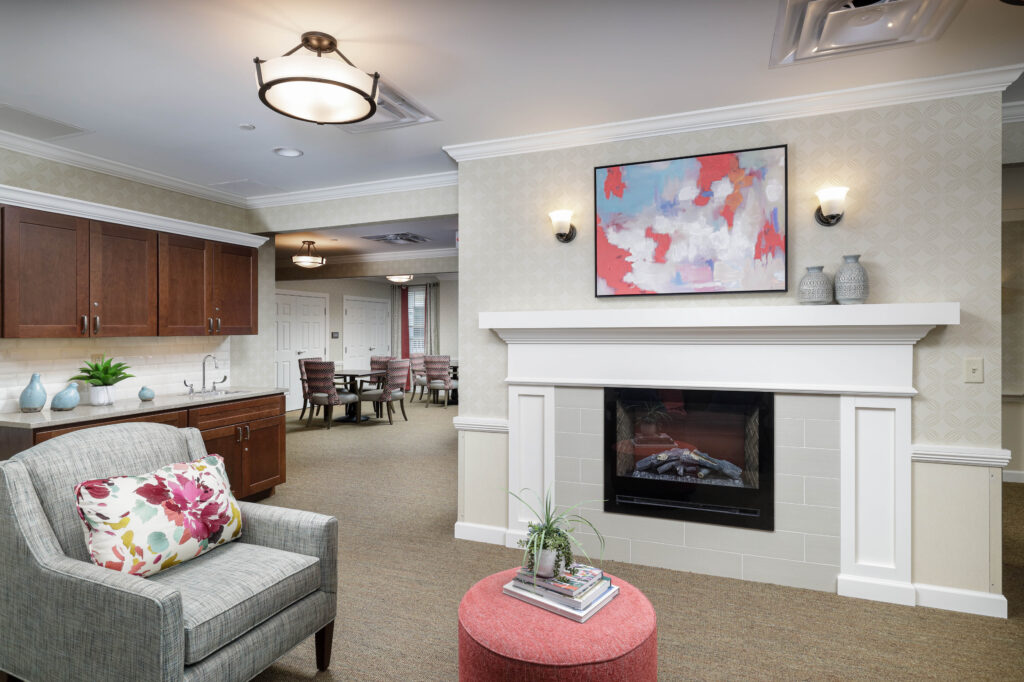A lounge area with a fireplace, abstract art, a grey armchair with a floral pillow, and a red ottoman, looking toward a dining area.