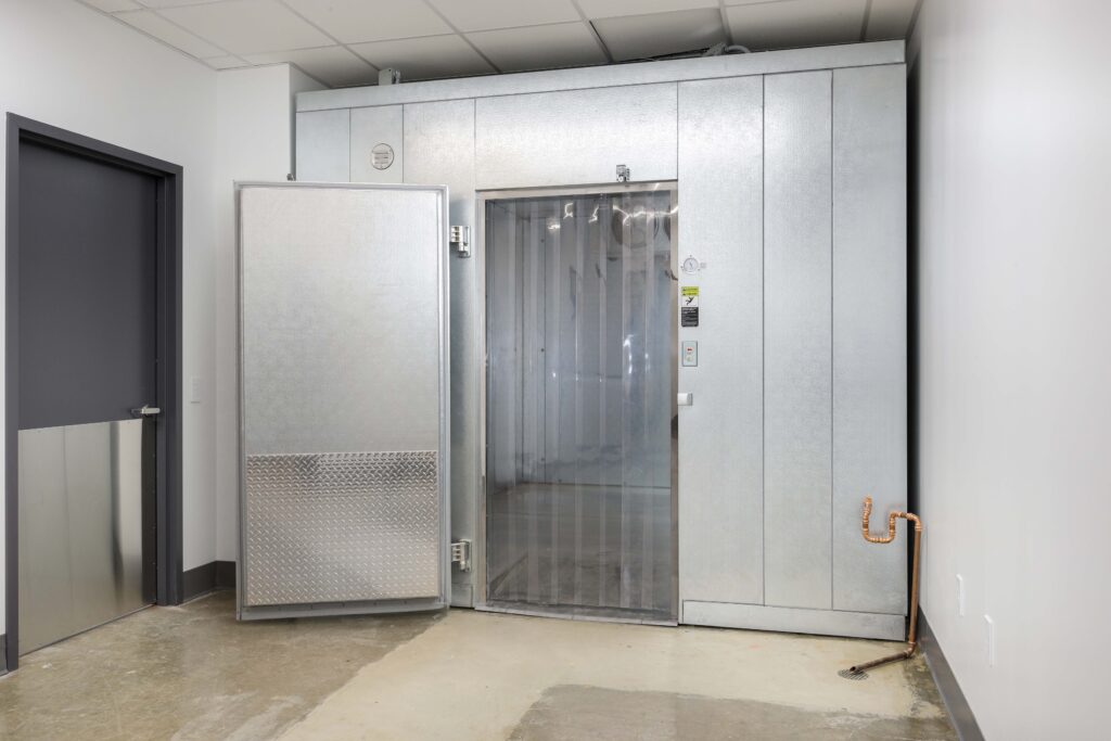 Large walk-in cooler with a heavy metal door standing open, revealing a plastic strip curtain entrance.