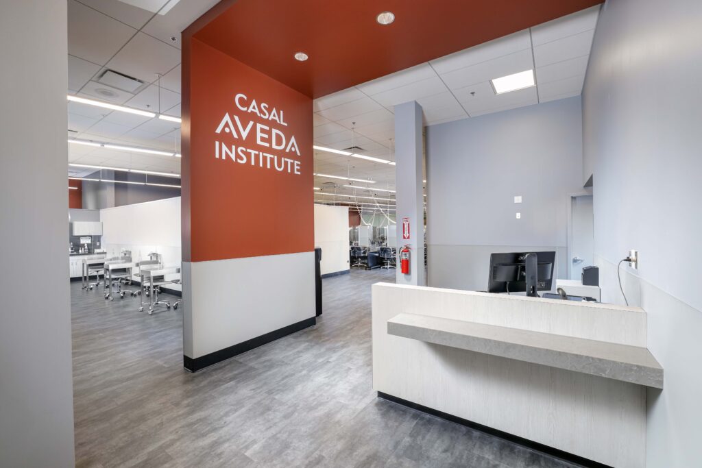 Interior view of the Casal Aveda Institute reception area, featuring a white front desk and an orange wall with the school’s name in white lettering.