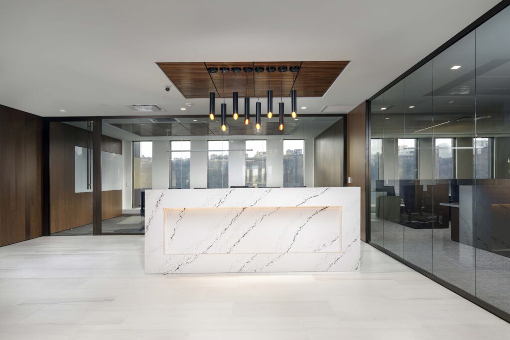 A marble reception desk sits in a modern office lobby under a row of hanging pendant lights, with glass-walled conference rooms in the background.