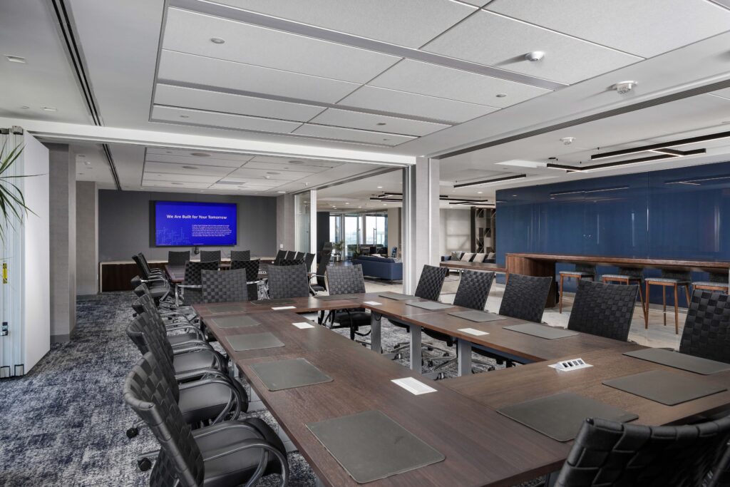 A conference room with a large U-shaped wooden table, black office chairs, and a monitor displaying text on a blue screen.