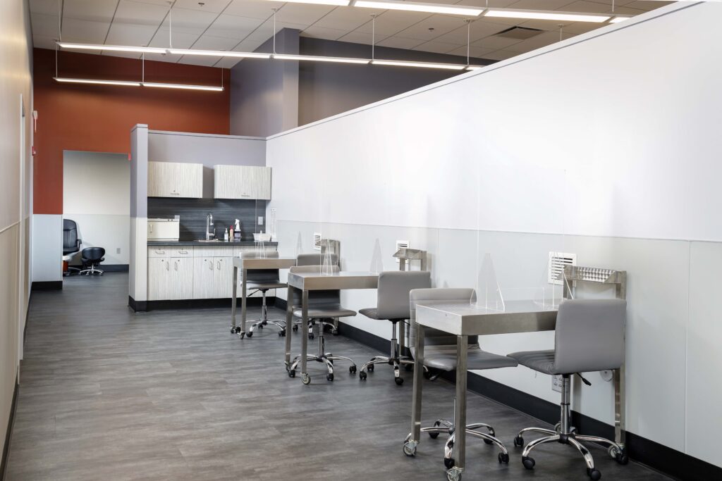 A room with rows of clinical metal tables with chairs and clear dividers, and a small kitchenette with cabinets and a sink along the back wall.