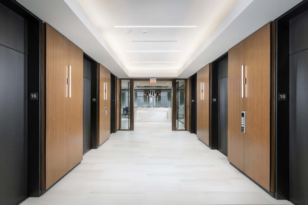 Elevator lobby with wood-paneled walls, floor numbers, and a reception desk visible through a glass doorway at the end of the hall.