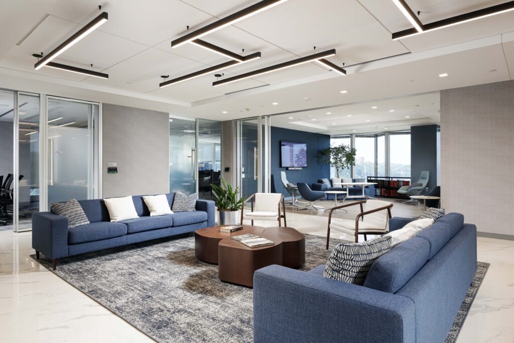 Modern office lounge area with two blue sofas, wooden coffee tables, and glass-walled meeting rooms in the background.
