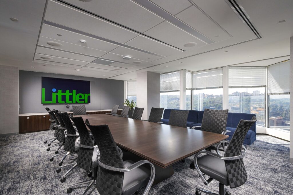 A conference room featuring a long dark wood table, several black office chairs, and a monitor displaying the Littler logo on the wall.