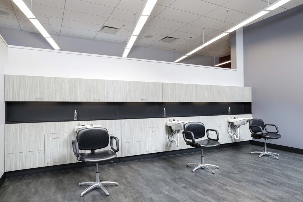 Three black salon chairs positioned in front of wall-mounted shampoo sinks and light-wood cabinetry.