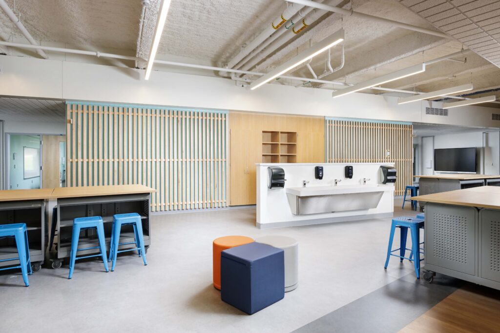 A classroom or lab space with blue stools, a central handwashing station, and a wood-slat feature wall.