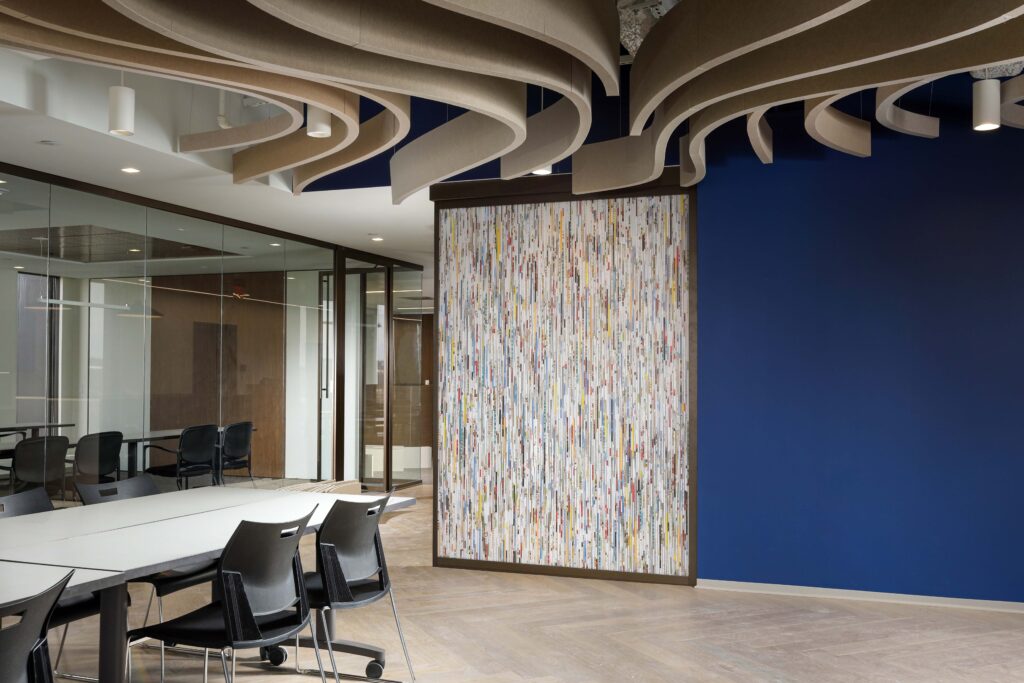 Modern conference room with a white table, chairs, a blue accent wall, and a decorative panel featuring vertical multicolored strips, beneath a sculptural, wave-patterned ceiling baffle system.