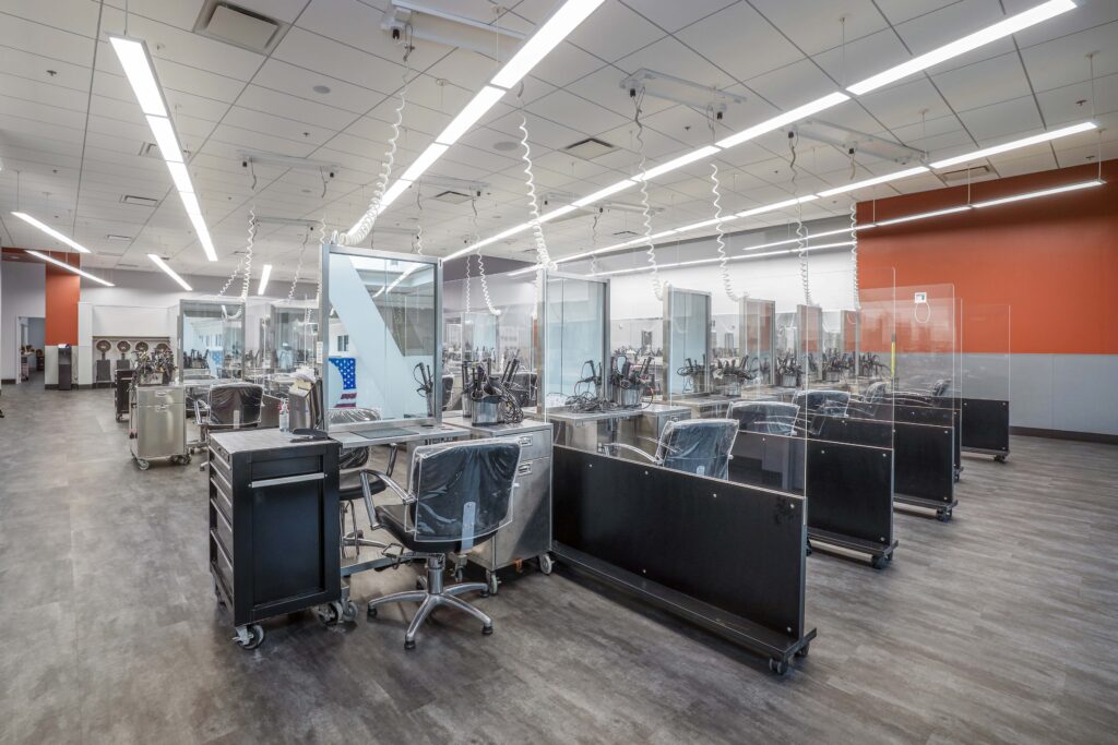 Rows of salon styling stations with chairs, equipment, and clear protective partitions on a grey floor.