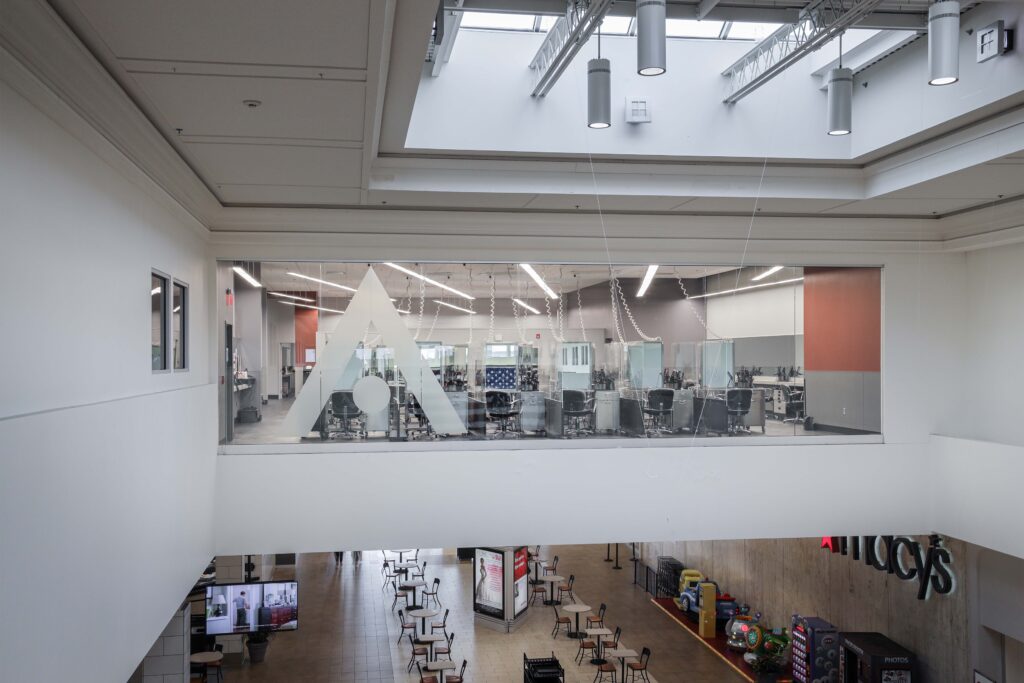 View from a balcony into an indoor shopping mall, showing a glass-walled office with a large triangular logo, a seating area with tables and chairs, and a Macy’s entrance.
