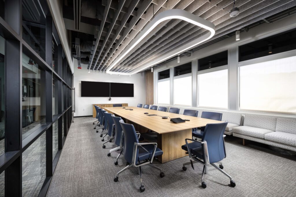 A modern conference room featuring a long wooden table, blue office chairs, and a large wall-mounted monitor under a minimalist rectangular light fixture.