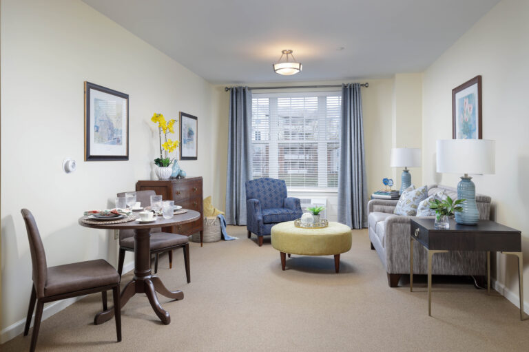 A furnished living room with a dining set, a blue patterned armchair, a yellow ottoman, and a grey sofa.