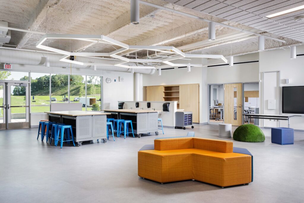 Modern classroom or meeting space with modular orange seating, desks with blue stools, and geometric hanging lights.