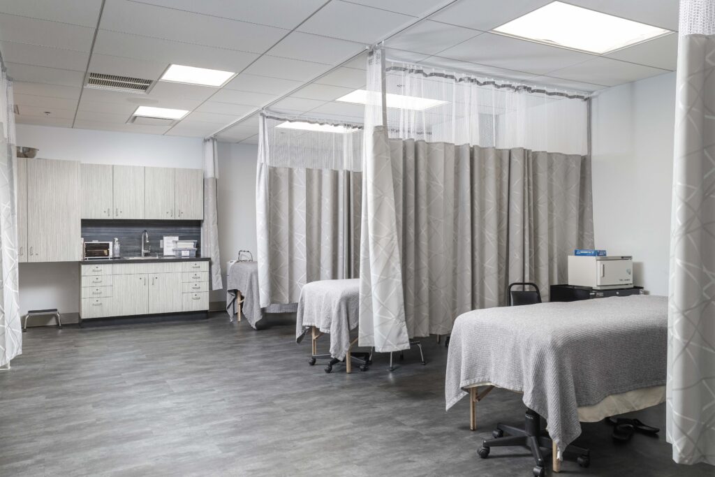 A clinical treatment room featuring multiple massage tables partitioned by privacy curtains, with a sink and cabinets along the back wall.