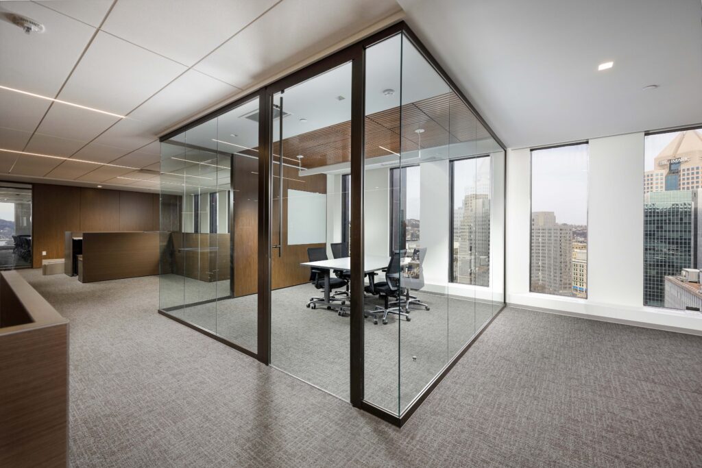 A glass-walled conference room with a white table and four chairs, situated in an office overlooking a city skyline.