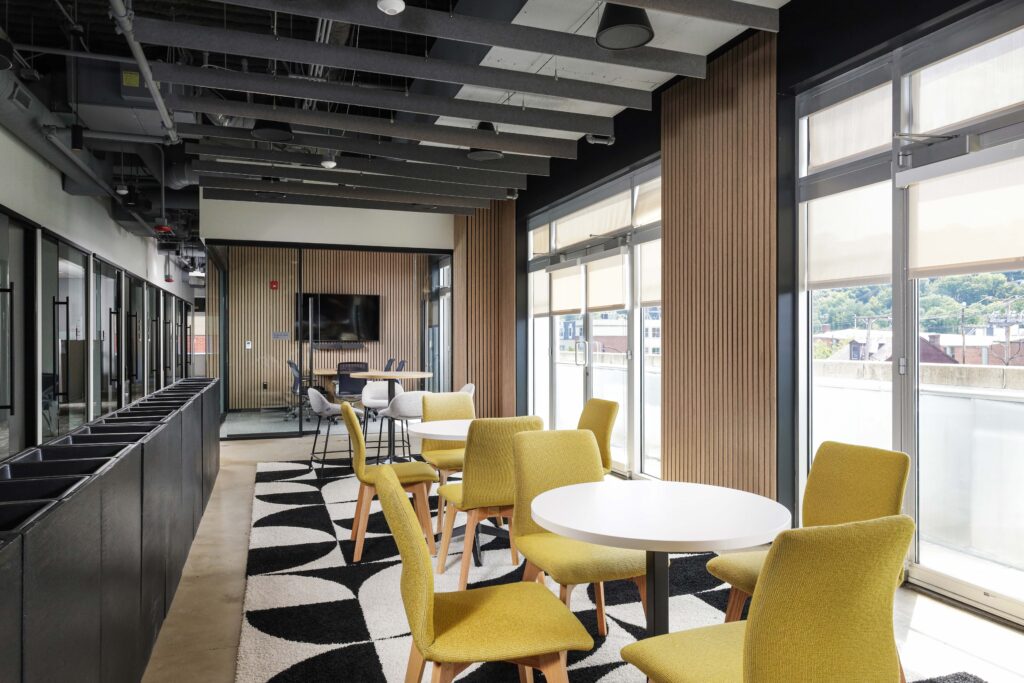 Modern office lounge featuring round white tables with yellow chairs, a geometric patterned rug, and floor-to-ceiling windows.
