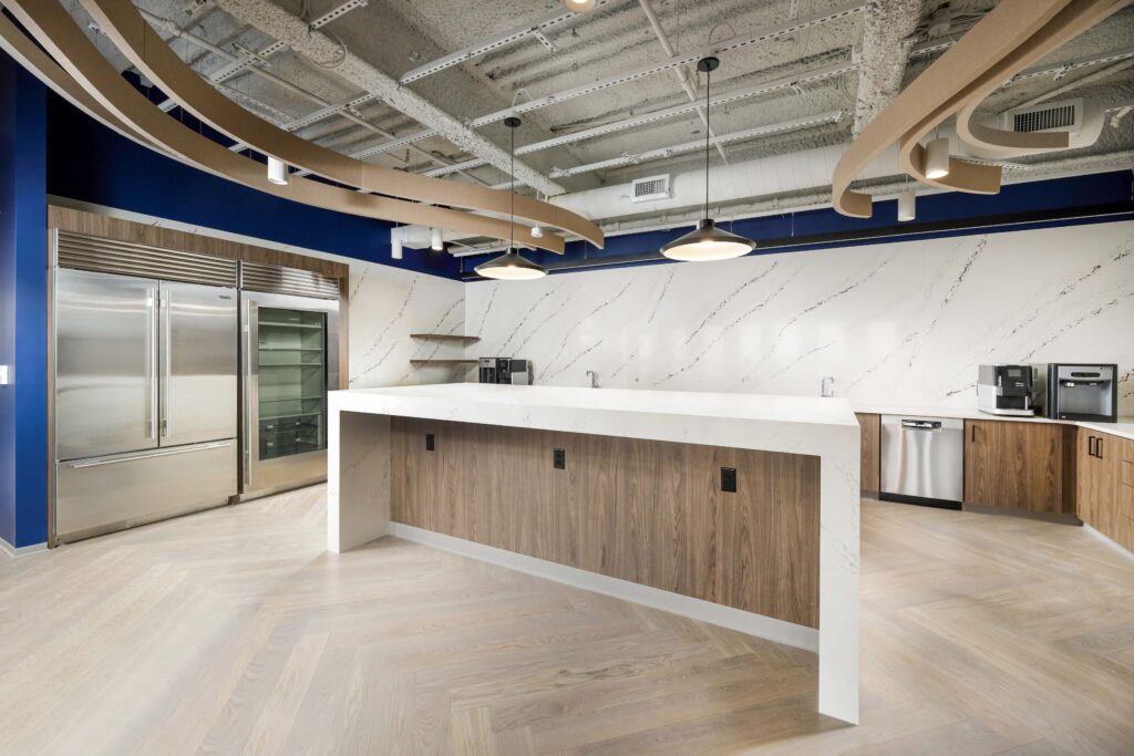 A modern office kitchen with a white marble waterfall island, stainless steel refrigerators, and wooden cabinetry.