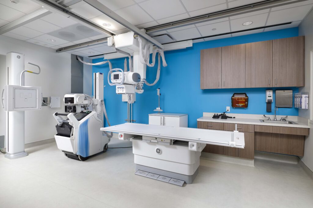 An X-ray examination room featuring a diagnostic table, a mobile X-ray unit, and wall-mounted imaging equipment.