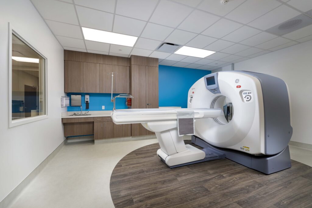A CT scanner in a medical imaging room with cabinets, a sink, and a blue accent wall.
