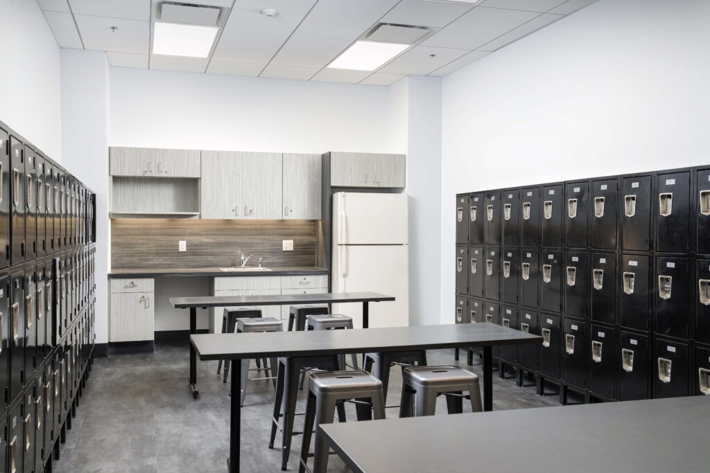 An office breakroom featuring two long tables with stools, a kitchenette with a white refrigerator, and walls lined with black lockers.