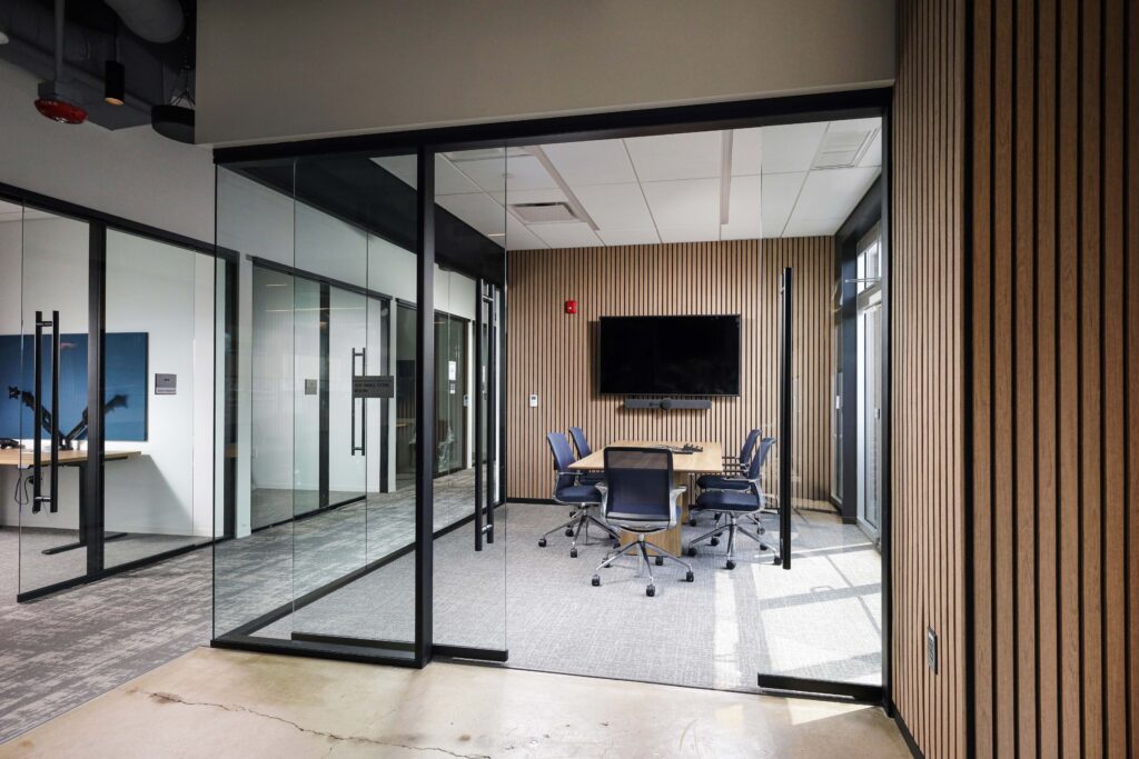 Glass-walled conference room with a wooden table, chairs, and a wall-mounted flat-screen television.
