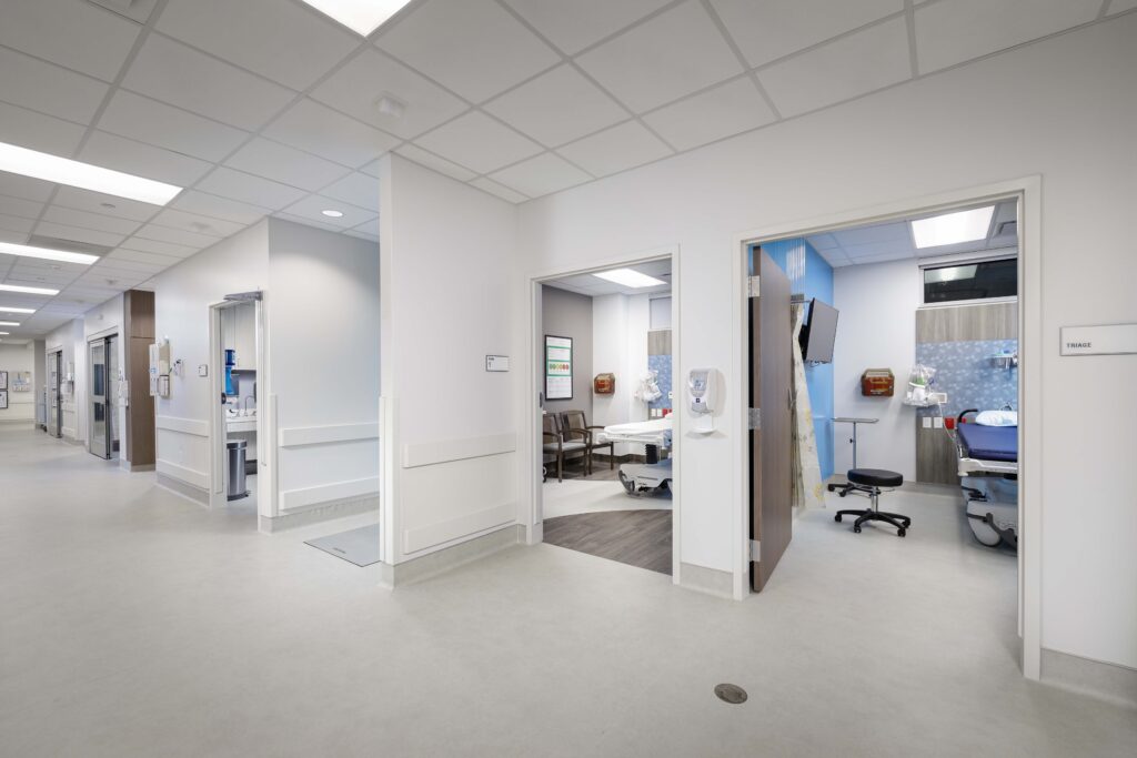 Hospital hallway showing open doorways into patient exam rooms equipped with medical beds and seating.