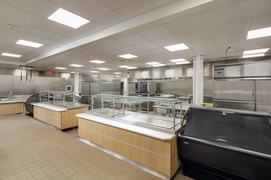 A commercial cafeteria kitchen featuring stainless steel prep stations, food service counters with sneeze guards, and industrial refrigerators.
