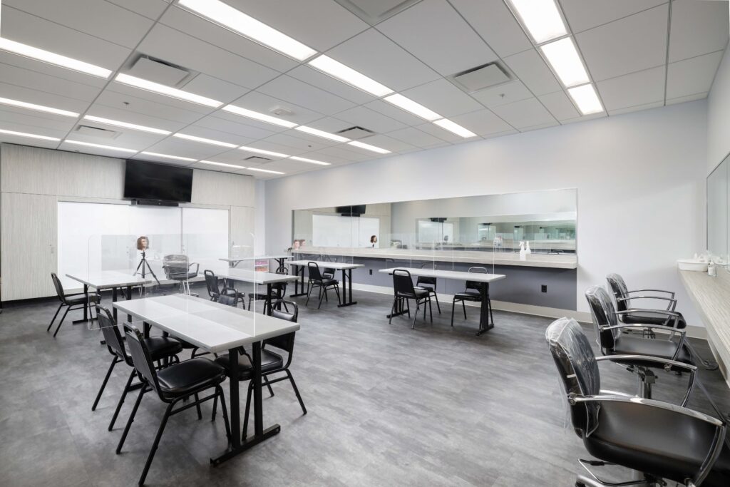A cosmetology training classroom with rows of tables, chairs, and mirrors, featuring clear protective screens between stations.