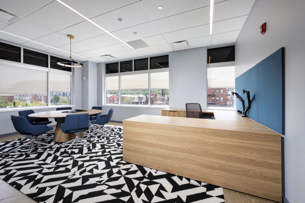 An office with a large L-shaped wood desk, a round meeting table with blue chairs, and a geometric black-and-white patterned rug.