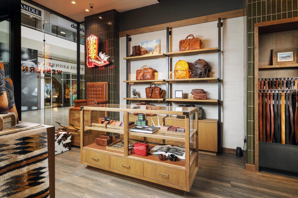 Interior of a western-style leather goods store with shelves displaying leather bags, wallets, and belts, featuring a glass display case and a neon sign shaped like a boot.