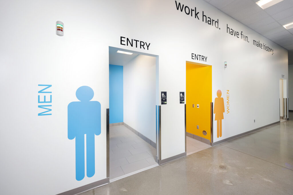 Two restroom entrances labeled “Men” in blue and “Women” in orange, located along a hallway with the text “work hard. have fun. make history.” on the wall.