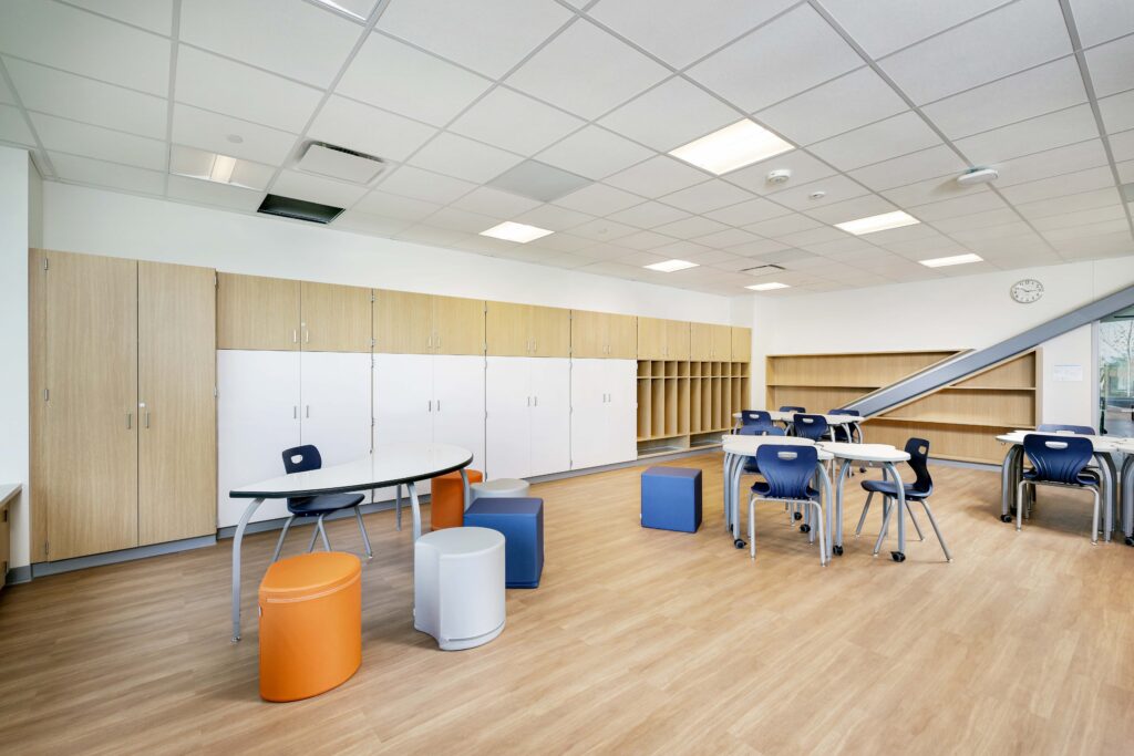 Modern classroom with light wood cabinets, white oval tables, blue chairs, and colorful round stools.