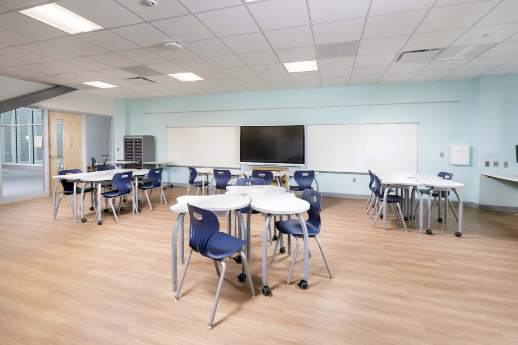 A classroom with light blue walls, wooden floors, and several clusters of white desks with blue chairs facing a central monitor and whiteboards.